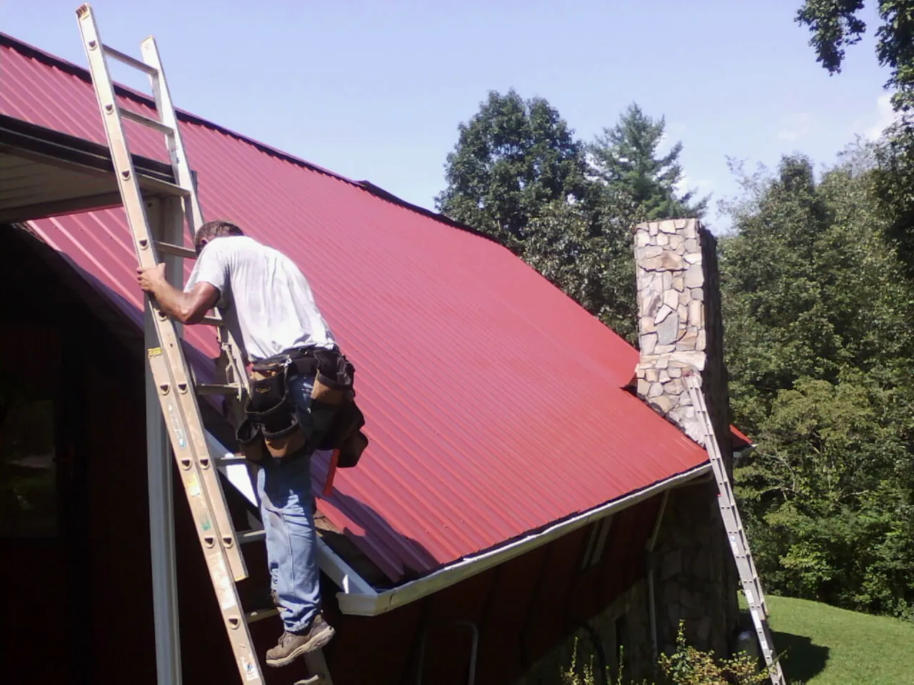Roofer on a ladder beside a red metal roof and stone chimney — Soffit & Fascia Repair by Roofing Brothers in Hill Afb